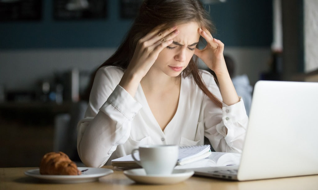 A person holds their temples while working on a laptop, with coffee and a notebook nearby. This scene shows fatigue, stress, and brain fog that can be linked to low-grade inflammation and gut health.