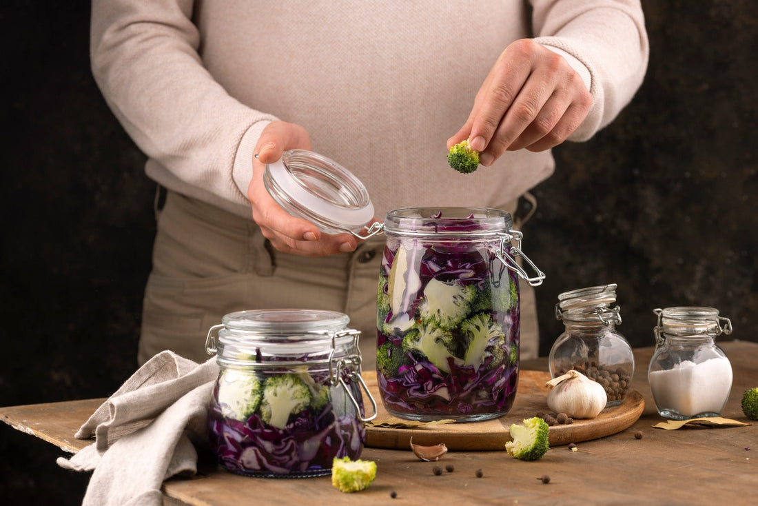 Person preparing fermented vegetables in glass jars, including cabbage and broccoli, as part of a traditional fermentation process.