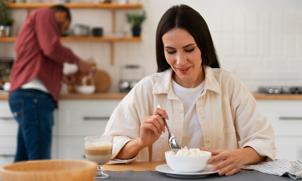 Woman eating yogurt at the breakfast table while another person prepares food in the kitchen, representing fermented foods, gut health, and healthy eating habits.