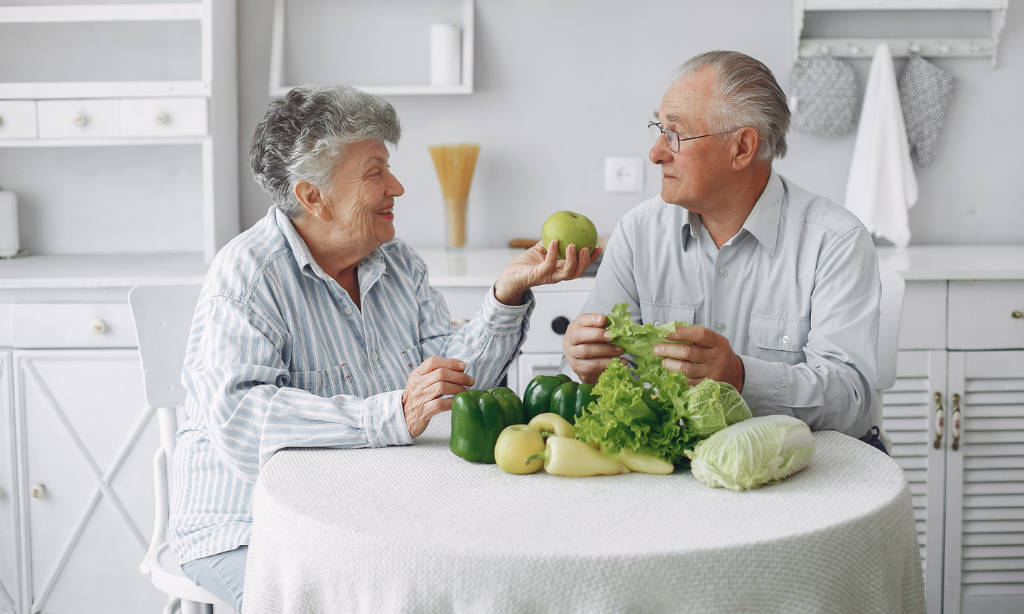 Elderly couple sitting at a table with fresh vegetables and fruit, discussing healthy eating at home.