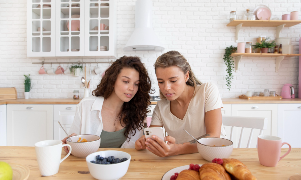 Two women sitting at a kitchen table eating breakfast, looking at a smartphone together with bowls of food, fruit, and pastries in front of them.