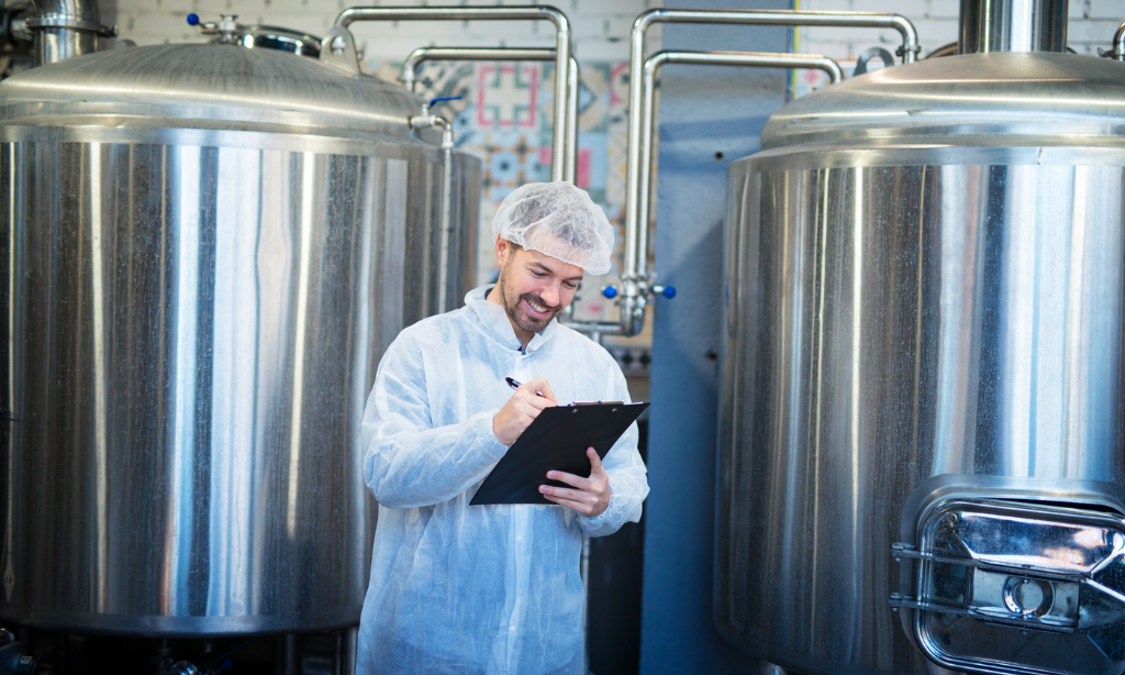 A supplement manufacturing worker wearing protective clothing and a hairnet stands between large stainless steel fermentation tanks while writing notes on a clipboard inside a clean production facility.