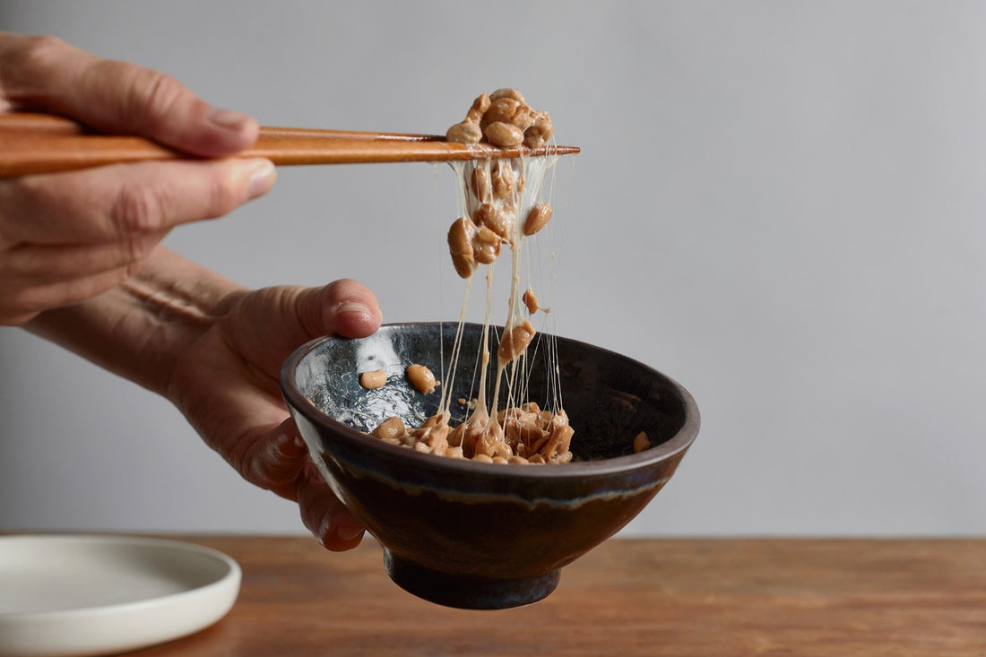 Hand holding chopsticks lifting sticky fermented natto soybeans from a black bowl on a wooden table.