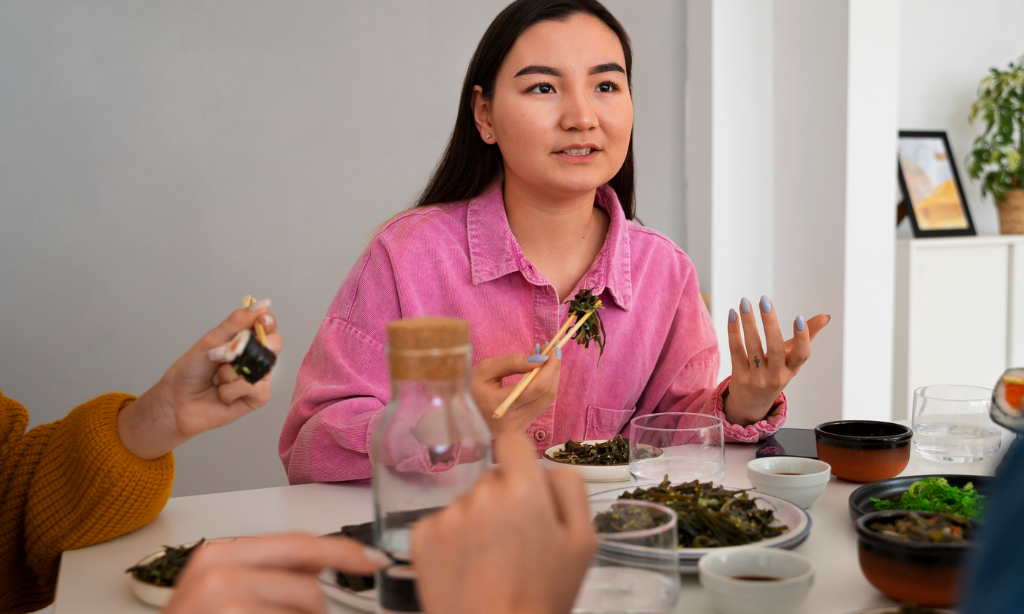 Woman in a pink shirt enjoying a meal with friends, discussing traditional foods around a dining table, highlighting growing global interest in Japanese longevity habits and mindful eating.
