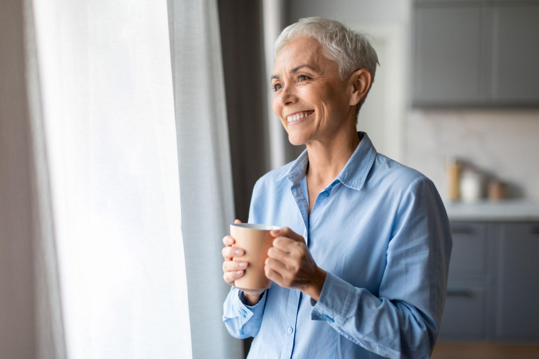 Older woman holding a cup and smiling by a window, symbolising healthy ageing and wellness.