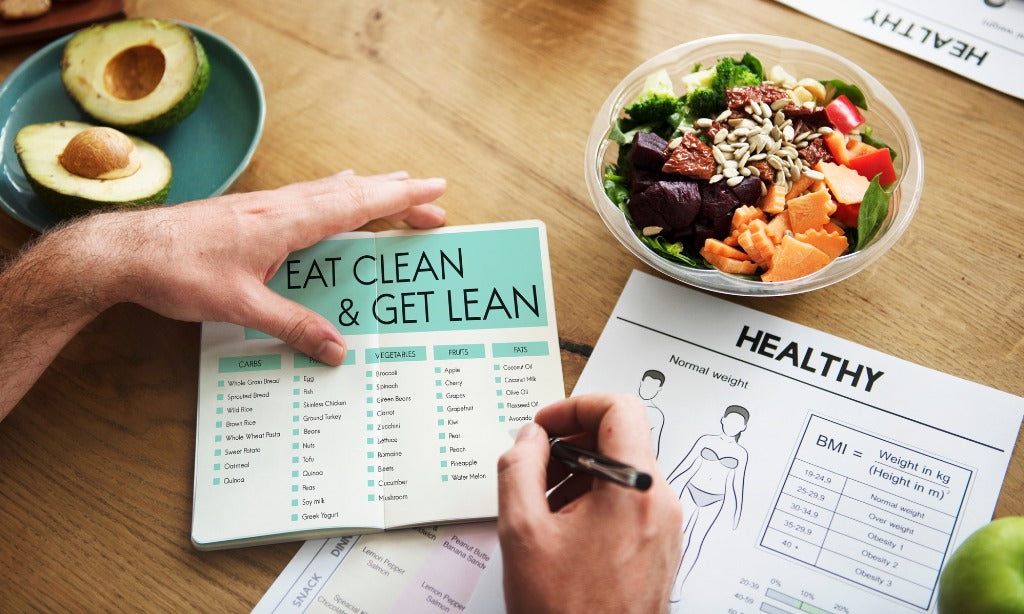 A person reviews weight and nutrition sheets on a desk beside a salad bowl and avocado, holding a booklet titled “Eat Clean & Get Lean,” with a BMI chart visible.