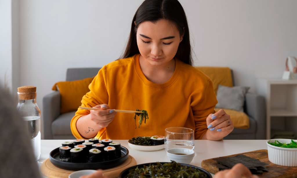 Young woman eating Japanese natto with chopsticks at a table with sushi and seaweed, highlighting traditional gut microbiome foods and the benefits of natto as a superfood.
