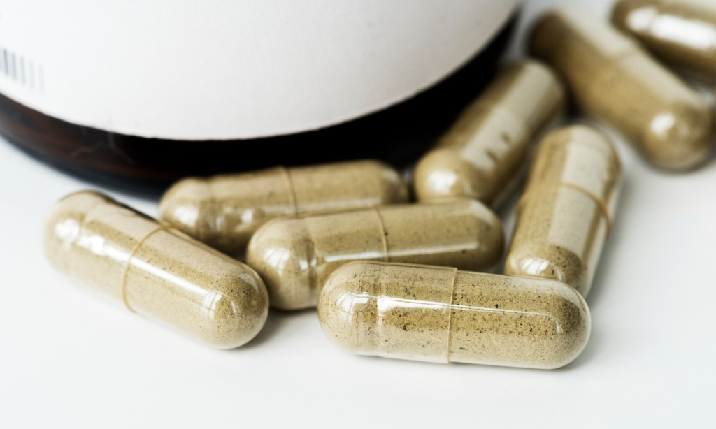 “Close-up of beige natto powder supplement capsules spilled from a white bottle on a white background.”