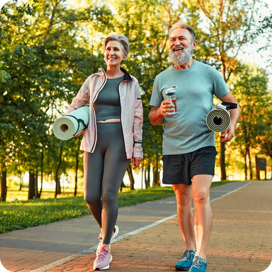 Man and woman walking outdoors on a path, holding yoga mats and drinks.